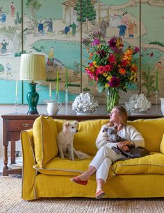 a woman sitting on a yellow couch with her dog in front of a wall painting