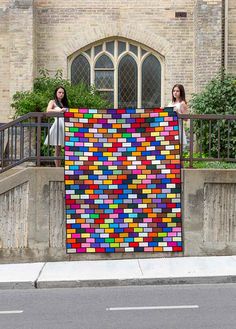 two women standing on a balcony holding up a large piece of art made out of colored blocks