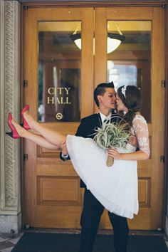 a bride and groom kissing in front of a city hall door with red high heels