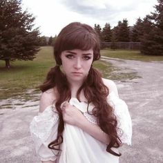 a woman with long hair wearing a white dress standing in the middle of a dirt road