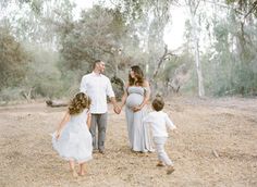 a pregnant woman and two children holding hands while walking through an open field with trees in the background