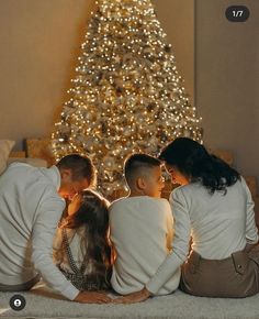 three people sitting on the floor in front of a christmas tree with lights all around them