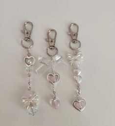 three key chains with charms attached to them on a white surface, one in the shape of a heart and two in the shape of a bow