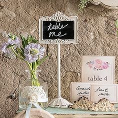 there is a table with flowers and cards on the shelf next to it, along with a chalkboard sign that says table me