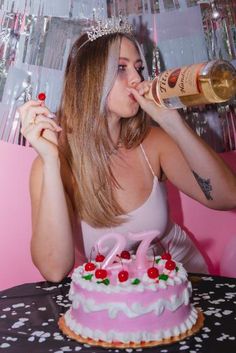 a woman sitting at a table drinking from a bottle next to a pink birthday cake