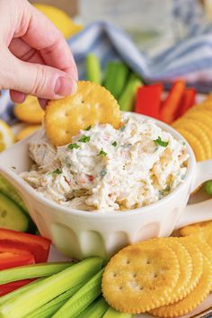 a hand dipping crackers into a bowl of dip surrounded by vegetables and crackers