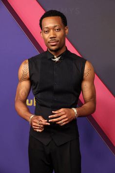 a man in black shirt and pants standing next to a purple wall with his hands on his hips