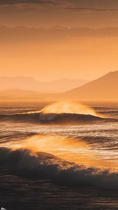 the sun is setting over the ocean with waves in the foreground and mountains in the background