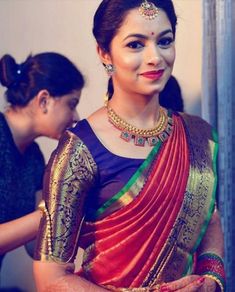 a woman in a red and green sari with gold jewelry on her neck, standing next to another woman