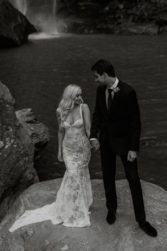 a man and woman standing next to each other in front of a waterfall holding hands