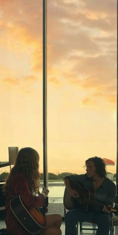 two people sitting on chairs playing guitars in front of a window with the sun setting