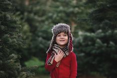 a young boy wearing a hat and scarf in front of pine trees with his hands on his chest