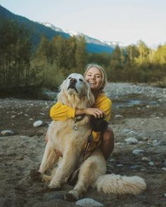 a woman sitting on the ground with her dog and taking a photo in front of mountains