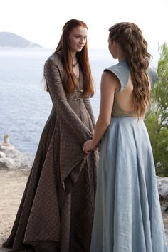 two young women dressed in medieval clothing standing next to each other near the water looking at each other