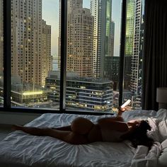 a naked woman laying on top of a bed in front of a window with tall buildings