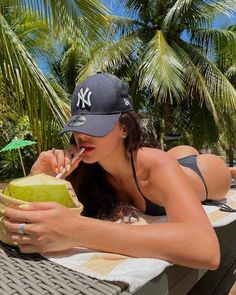 a woman laying on top of a table next to a green coconut and drinking from a straw