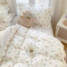 a bed with white sheets and pink flowers on it, next to a basket full of pillows