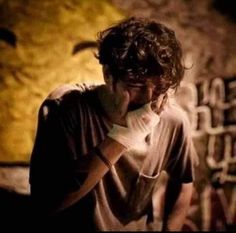 a man holding his hand to his face while standing in front of a wall with writing on it