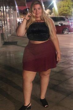 A woman poses confidently on a city sidewalk at night, wearing a black crop top, burgundy skirt, and black sandals. Leather Skirt, Leather