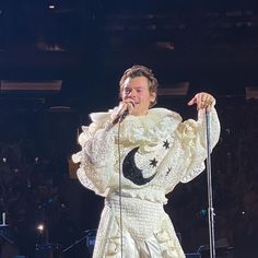 a man in a white dress singing into a microphone on stage with an audience behind him