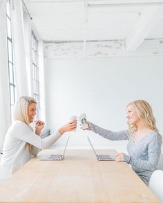 two women sitting at a table with laptops in their hands and one holding a coffee mug