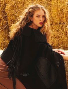 a woman sitting on top of hay bales wearing a black coat and brown pants