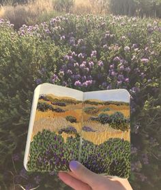 a hand holding an open book in front of some purple and yellow flowers on the ground