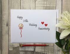 an anniversary card with a red pen next to it and a white flower on a wooden table