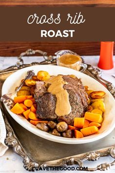 a roast with gravy and roasted vegetables in a white bowl on a silver platter