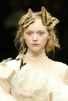 a woman with two snakes on her head and one snake on her shoulder, walking down the runway