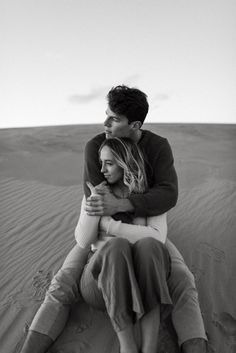 a man and woman sitting on top of a sand dune in the desert, hugging