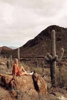 a woman sitting on top of a large rock next to a tall saguada