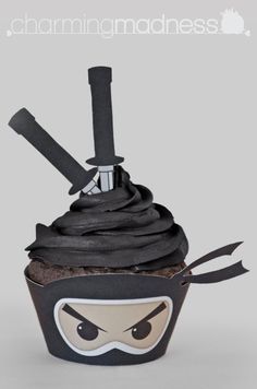 cupcake with ninja face and swords on top that says charming madness, it's chocolate frosting