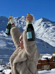 a woman holding two bottles of champagne in the snow with mountains in the back ground