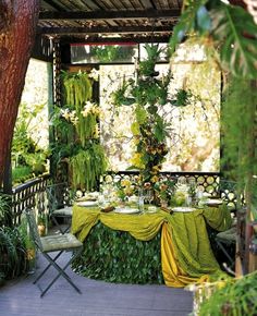 an outdoor dining area with table and chairs covered in green cloths, surrounded by greenery