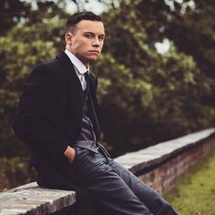 a man in a suit and tie sitting on a stone wall with trees in the background