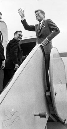 two men in suits wave from an airplane door as another man stands on the stairs