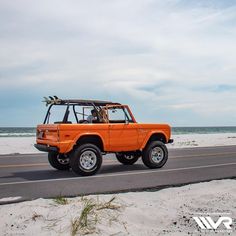 an orange jeep driving down the road by the beach with surfboards on top and two people in the back