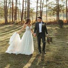 a bride and groom holding hands walking through the grass in front of some tall trees