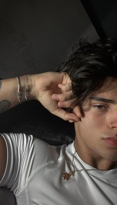 a young man with tattoos on his arm and hand resting against his head, wearing a white t - shirt