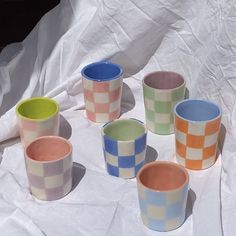 six cups sitting on top of a white sheet covered in plastic wrappers and one cup has a green, blue, orange checkerboard pattern