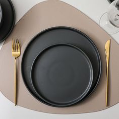 a table setting with black plates and gold cutlery on the placemat, wine glasses and utensils