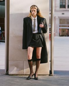 a woman in short skirt and coat leaning against a wall with headphones on her ears