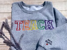 Custom embroidery in sweatshirt style! Please send a message before you order with custom embroidery requests outside of what is standard on the sweatshirt.  Featured sweatshirt is in the color sports grey with "TEACH" in red, orange, yellow, green and purple. The sleeve is embroidered with your prefix and last name in black with an apple underneath. * 8 oz. 50/50 US cotton/polyester * Classic fit * Unisex * 1x1 rib with spandex * 2-end midweight fleece fabric * Double-needle stitching on should Teacher Embroidery Designs Shirts, Embroidered Sports Sweatshirt, School Sweatshirt With Embroidered Text And Crew Neck, School Crew Neck Sweatshirt With Embroidered Text, School Crew Neck Sweatshirt With Embroidered Logo, Crew Neck Sweatshirt With Embroidered Graphics For School, Graphic Embroidered Crew Neck Sweatshirt For School, Embroidered Crew Neck Sweatshirt For School, Multicolor Embroidered Text Crew Neck Sweatshirt