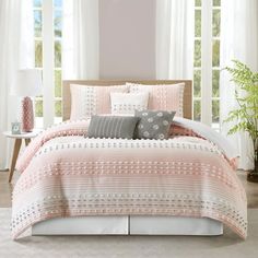 a bed with pink and white comforters in front of two windows next to a potted plant