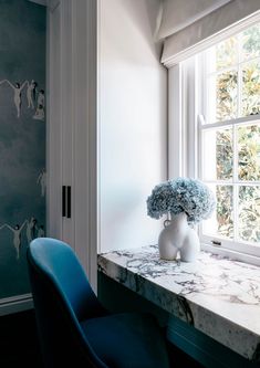 a blue chair sitting in front of a window next to a white vase filled with flowers