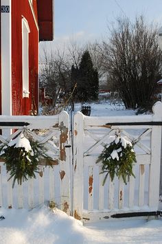 a white gate with wreaths on it in front of a red house covered in snow