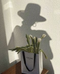 a white bag with flowers in it sitting on a table next to a shadow of a man's hat