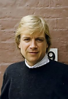 a woman with blonde hair wearing a black sweater and white collared shirt standing in front of a brick wall