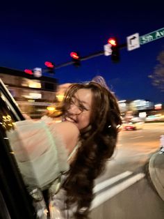 a woman leaning out the window of a car on a city street at night with traffic lights in the background